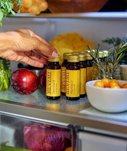 A hand reaching for bottles of Bevimi Uno Protect chilling on a refrigerator shelf beside fresh produce.