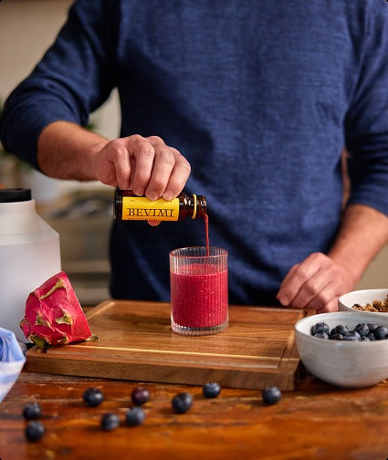 A person pouring a bottle of Bevimi Uno Protect into a glass of a red beverage at a kitchen counter.